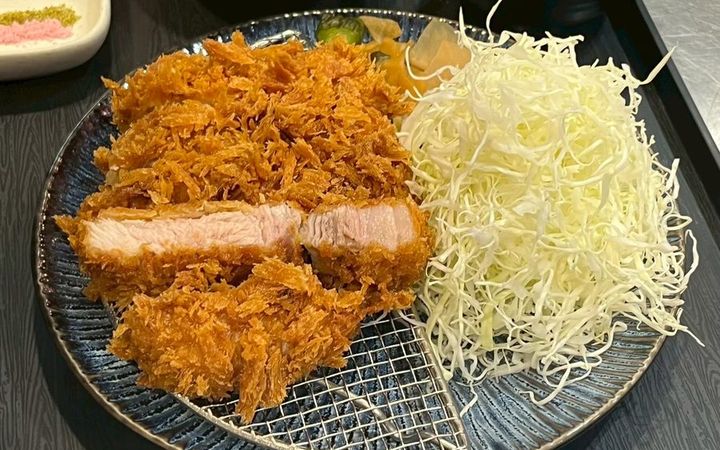 ทงคัตสึเกล็ดขนมปังฟูหั่นให้เห็นเนื้อสีชมพูอ่อน วางบนตะแกรงในจานสีน้ำเงิน ข้างกองกะหล่ำปลีซอยเส้นฝอยที่ตั้งสูง